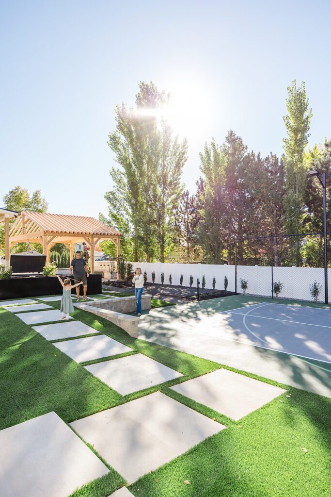 Back exterior of a Utah custom home with sport court, patio space, and beautiful yard design